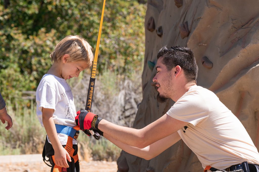 A young child wearing a harness stands next to a rock climbing wall, part of an engaging outdoor education program. An adult, wearing gloves, helps adjust the child's harness. Both are outdoors on a sunny day with trees in the background.