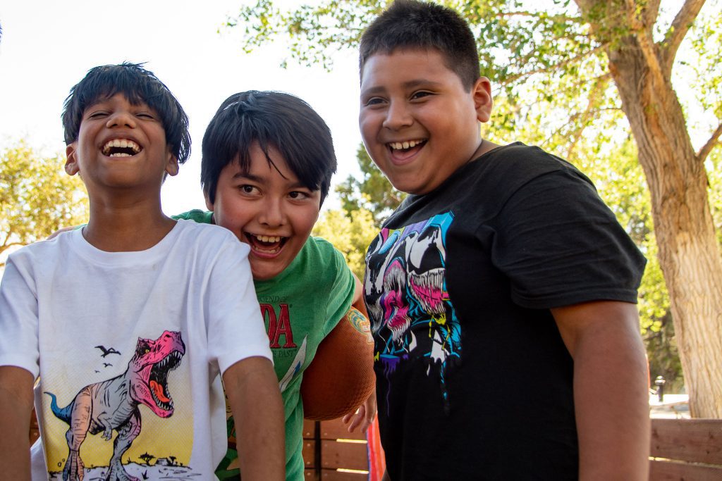 Three boys are smiling and laughing outdoors, standing close together. One is holding a football, embodying the spirit of outdoor fun. They are wearing casual t-shirts featuring colorful graphics, with trees in the background suggesting a park setting perfect for informal outdoor education.