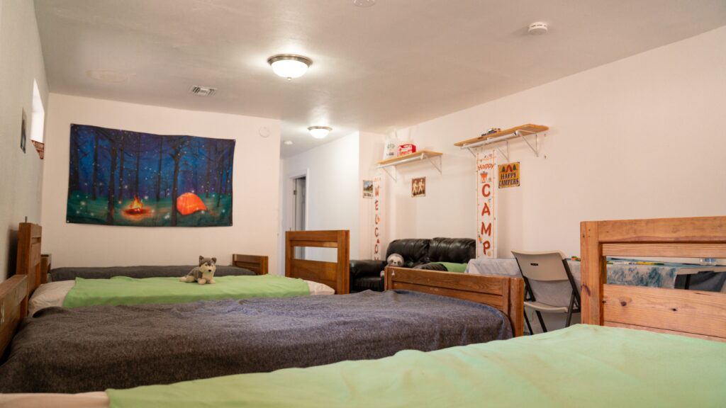 A cozy room with three single beds, each with green blankets, wooden frames, and plush toys. There is a couch, a desk with a chair, wall shelves, and camping-themed decorations on the white walls.