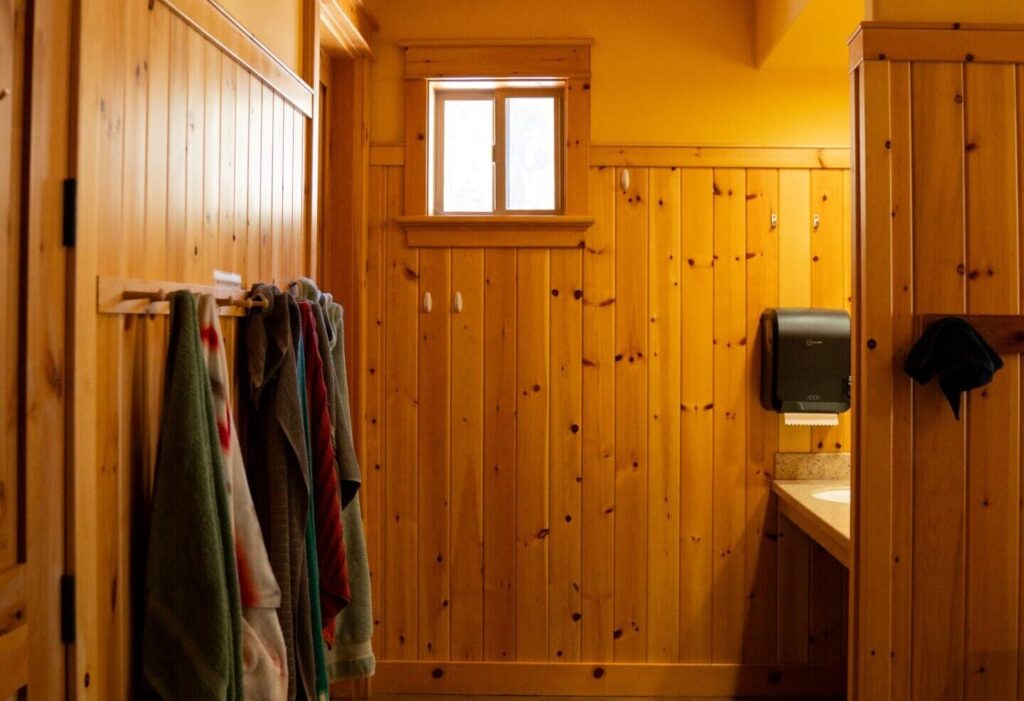 A warm, wooden-paneled restroom with several colorful towels hanging on hooks, a small window letting in natural light, and a sink area with a soap dispenser.