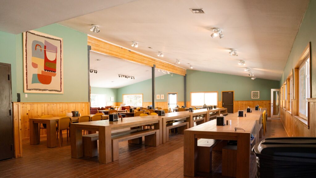 A spacious dining hall with wooden tables and benches, green and wood-paneled walls, modern art on the wall, and large windows letting in natural light. Napkin holders sit on the tables.
