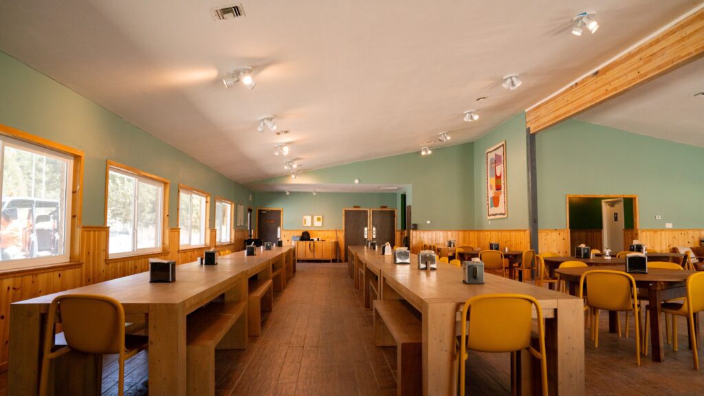 A spacious dining hall with long wooden tables, yellow chairs, large windows on the left, and artwork on the light green and wood-paneled walls. Natural light fills the room, creating a bright, inviting atmosphere.