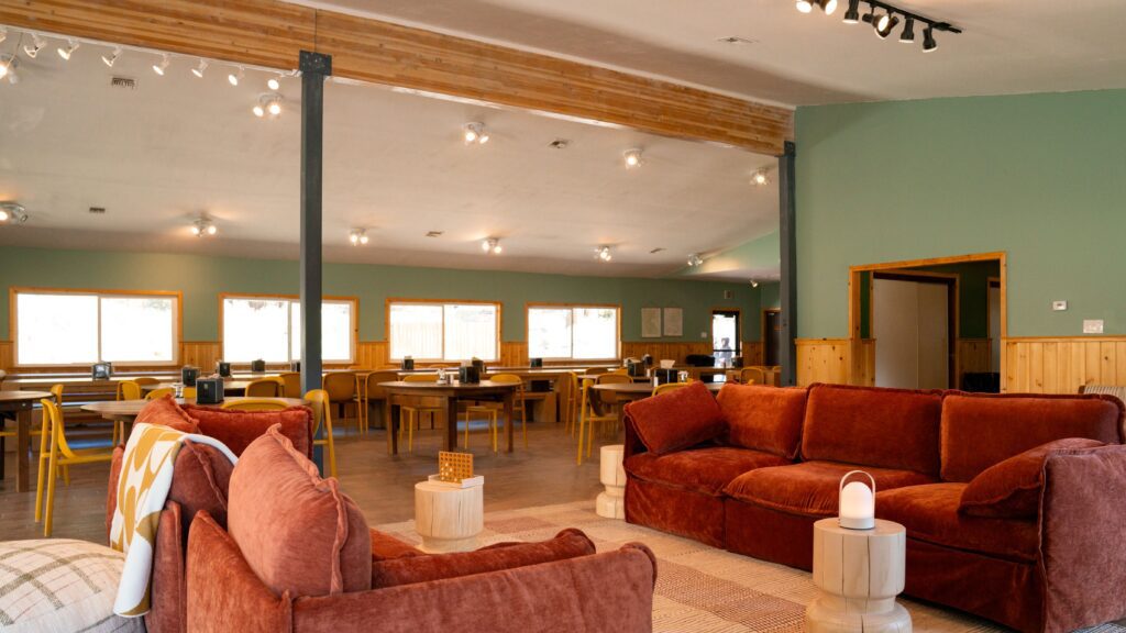 A spacious room with green walls features several wooden tables and chairs in the background and two rust-colored sofas with small round tables in the foreground. Large windows let in natural light.