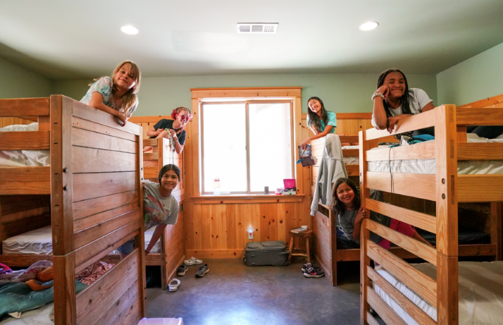Six girls smile and pose on wooden bunk beds in a bright, cozy cabin room with a window in the center, light green walls, and scattered luggage on the floor.